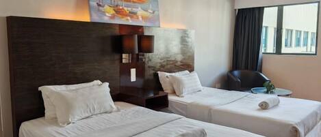 A hotel room with two beds, a painting above the headboard, a desk with a chair, and a window with curtains.