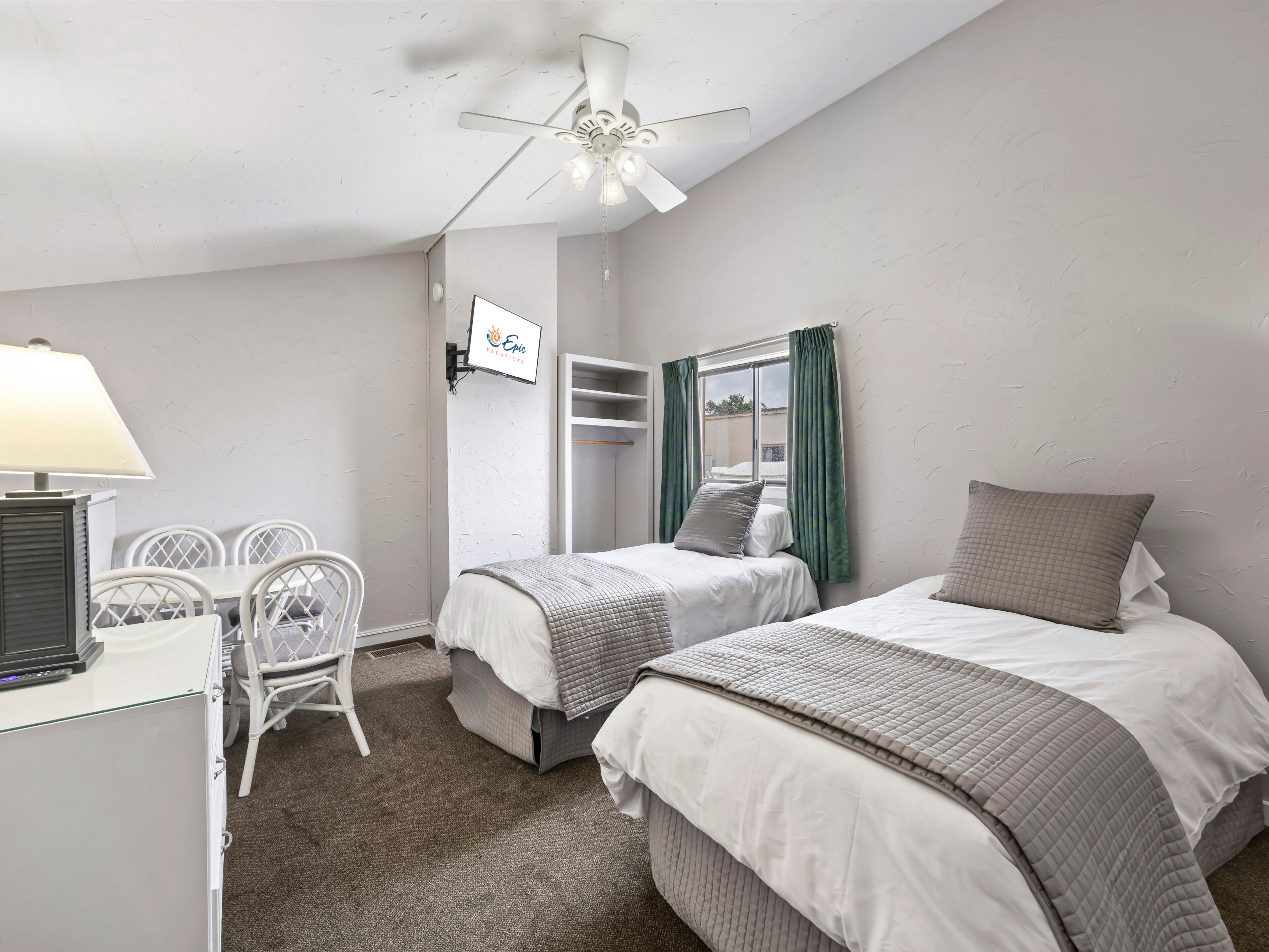 A hotel room with two beds, a desk with a lamp, a chair, a wardrobe, and a ceiling fan.