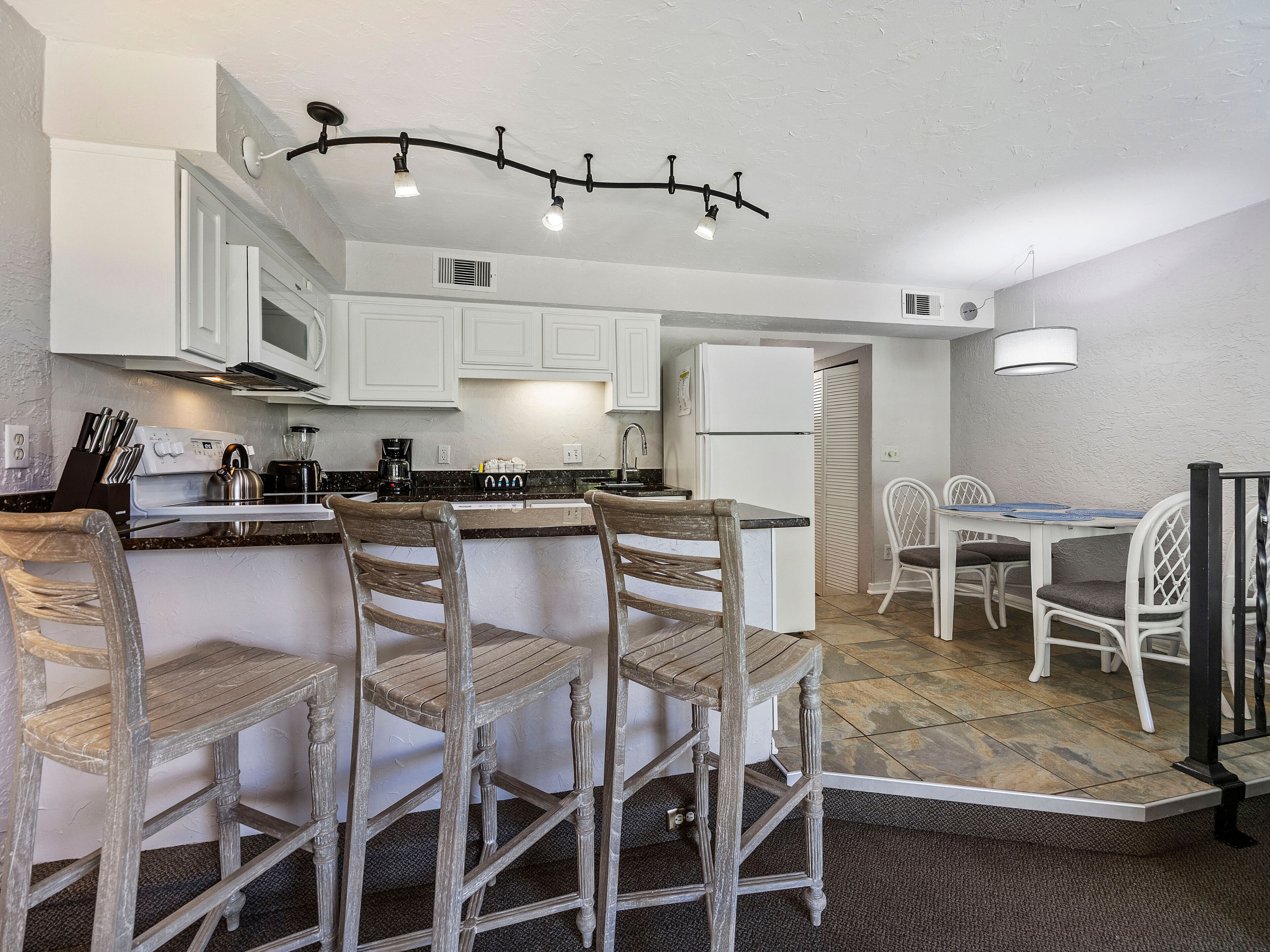 A kitchen with white cabinets, a central island with bar stools, a refrigerator, and a dining area with a table and chairs.