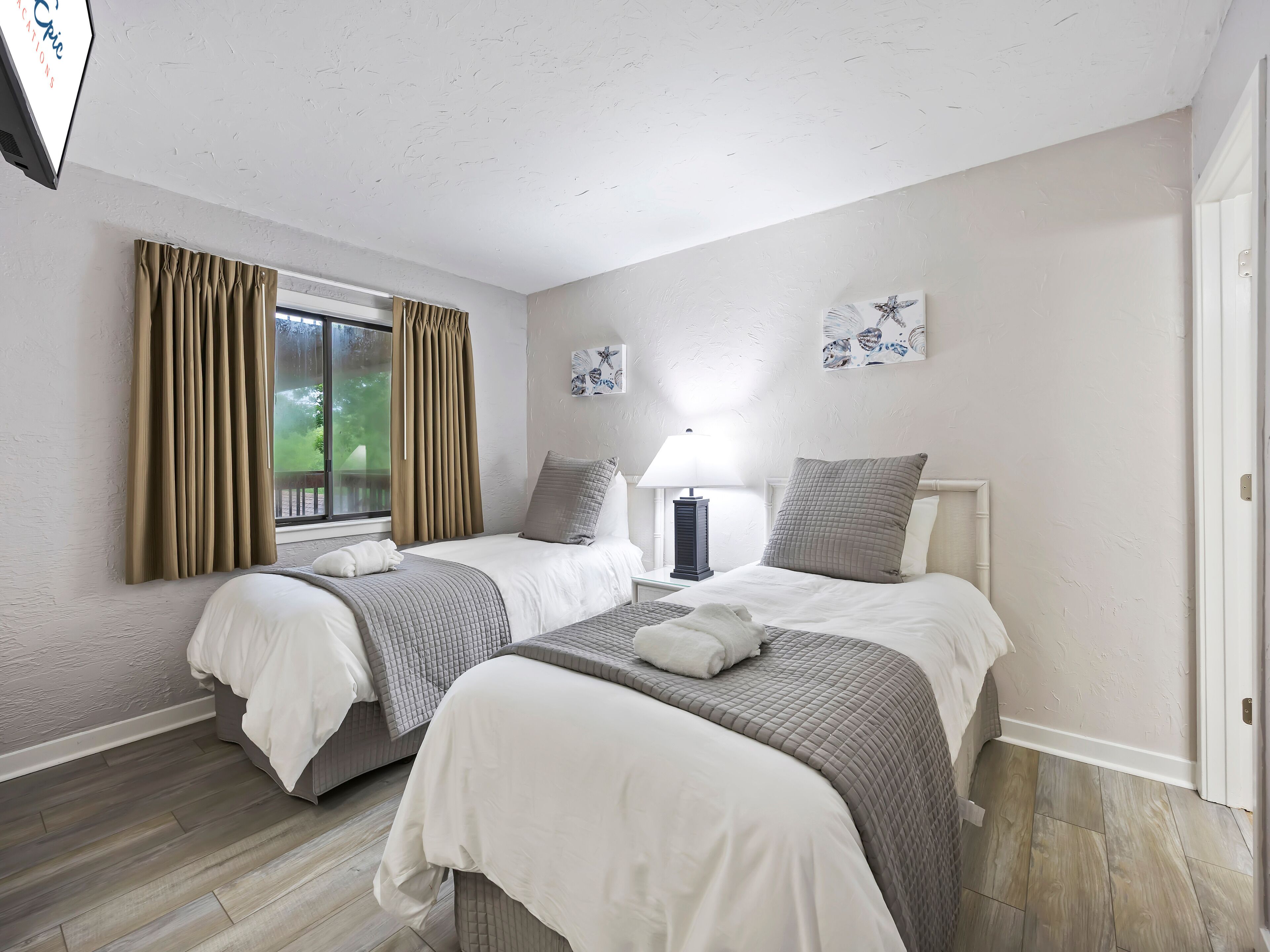 A hotel room with two beds, a window with curtains, a nightstand with a lamp, and artwork on the wall.