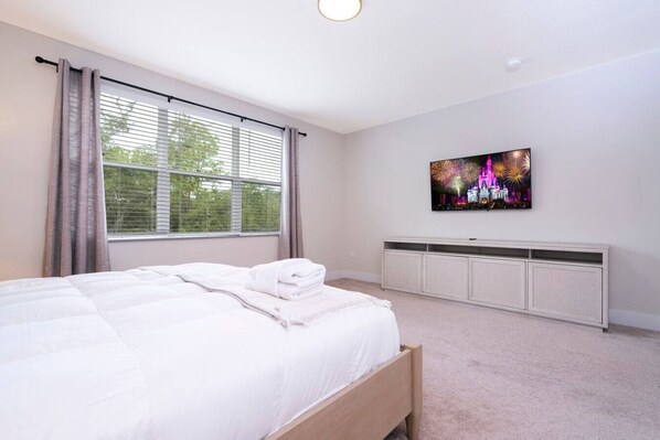 A bedroom with a bed, a window with blinds, a white dresser, and a wall-mounted TV.