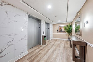 Un pasillo con piso de mármol, dos ascensores, un contenedor de reciclaje y un banco de madera.