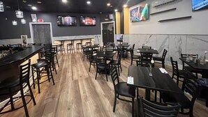 A restaurant with multiple tables and chairs, a bar area, and two TVs mounted on the wall.