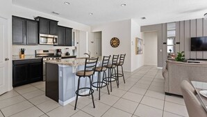 A modern kitchen with dark cabinets, a central island with bar stools, and a dining area with a TV and seating. - Entire House Davenport ~ Pool ~ Resort Access (Davenport)