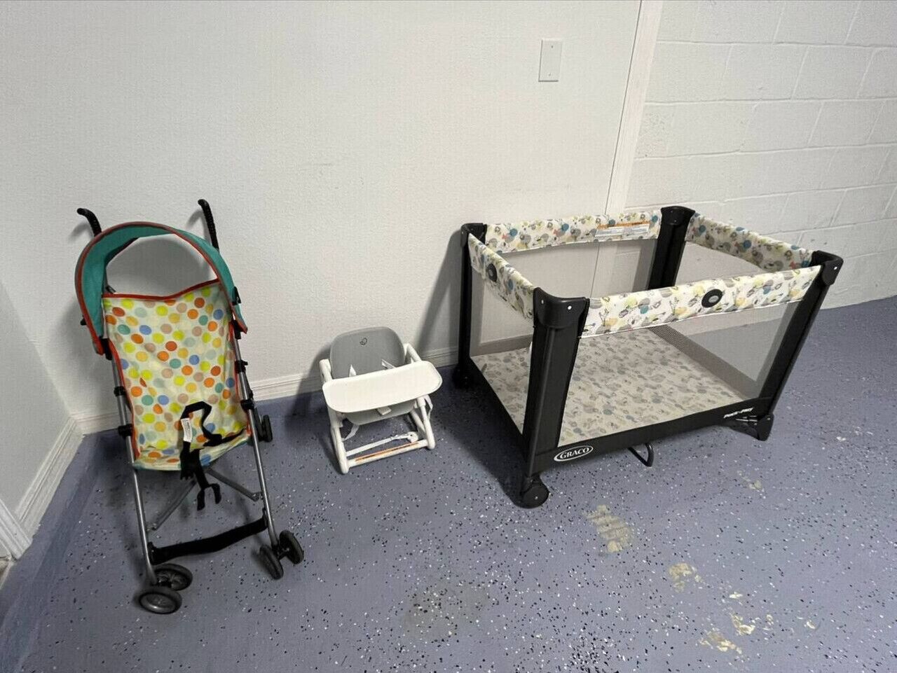 A baby playpen and a stroller in a room with a plain white wall and a speckled floor.