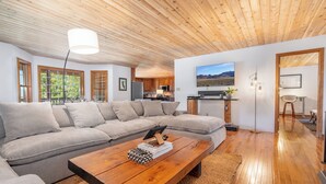 A modern living room with a wooden ceiling, a large sofa, a coffee table, and a kitchen in the background. - Reverie Ranch @ Gold Mountain 136 Acre w/ Lakeview, Sauna, & Nat'l Forest Access (Milford)