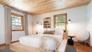 A bedroom with a wooden ceiling, a bed with white bedding, two bedside lamps, a round table with a lamp, and a view of trees through the windows. - Reverie Ranch @ Gold Mountain 136 Acre w/ Lakeview, Sauna, & Nat'l Forest Access (Milford)