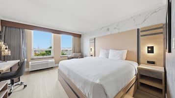A hotel room with a large bed, a desk, a chair, and a view of the cityscape through the window.
