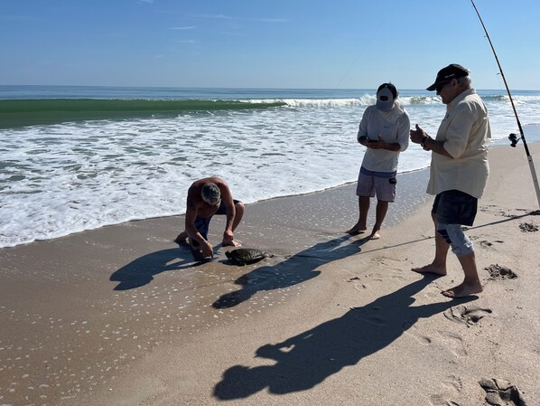 A beach scene with three people, one crouching near the water's edge, another standing with a fishing rod, and a third observing. - Surfside Suites (Cocoa Beach)