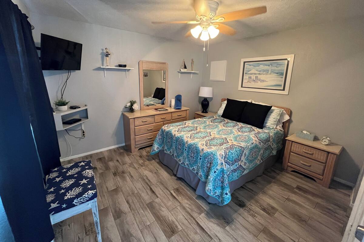 A bedroom with a bed, dresser, nightstands, a ceiling fan, a TV, and a painting on the wall.