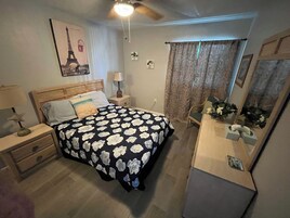 Un dormitorio con cama, mesitas de noche, cómoda, ventilador de techo, espejo y ventana con cortinas.