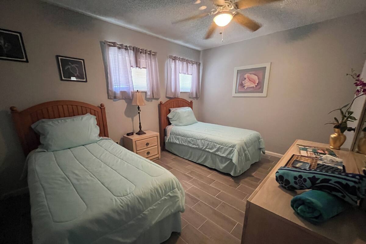 A room with two single beds, a nightstand with a lamp, a ceiling fan, and a small table with a vase and a plant.