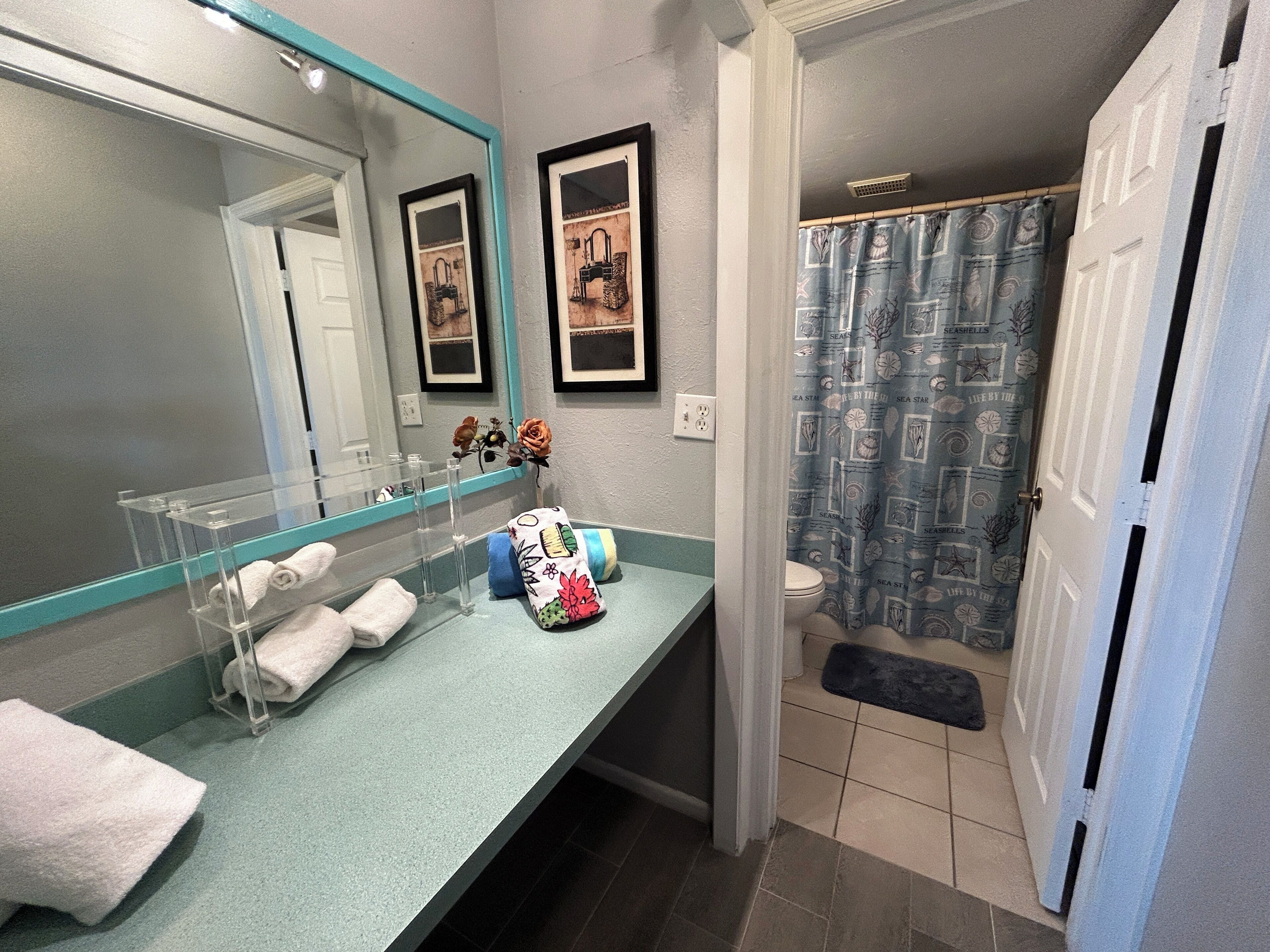 A bathroom with a teal countertop, a large mirror, two framed pictures, a toilet, and a shower curtain with a sea theme.