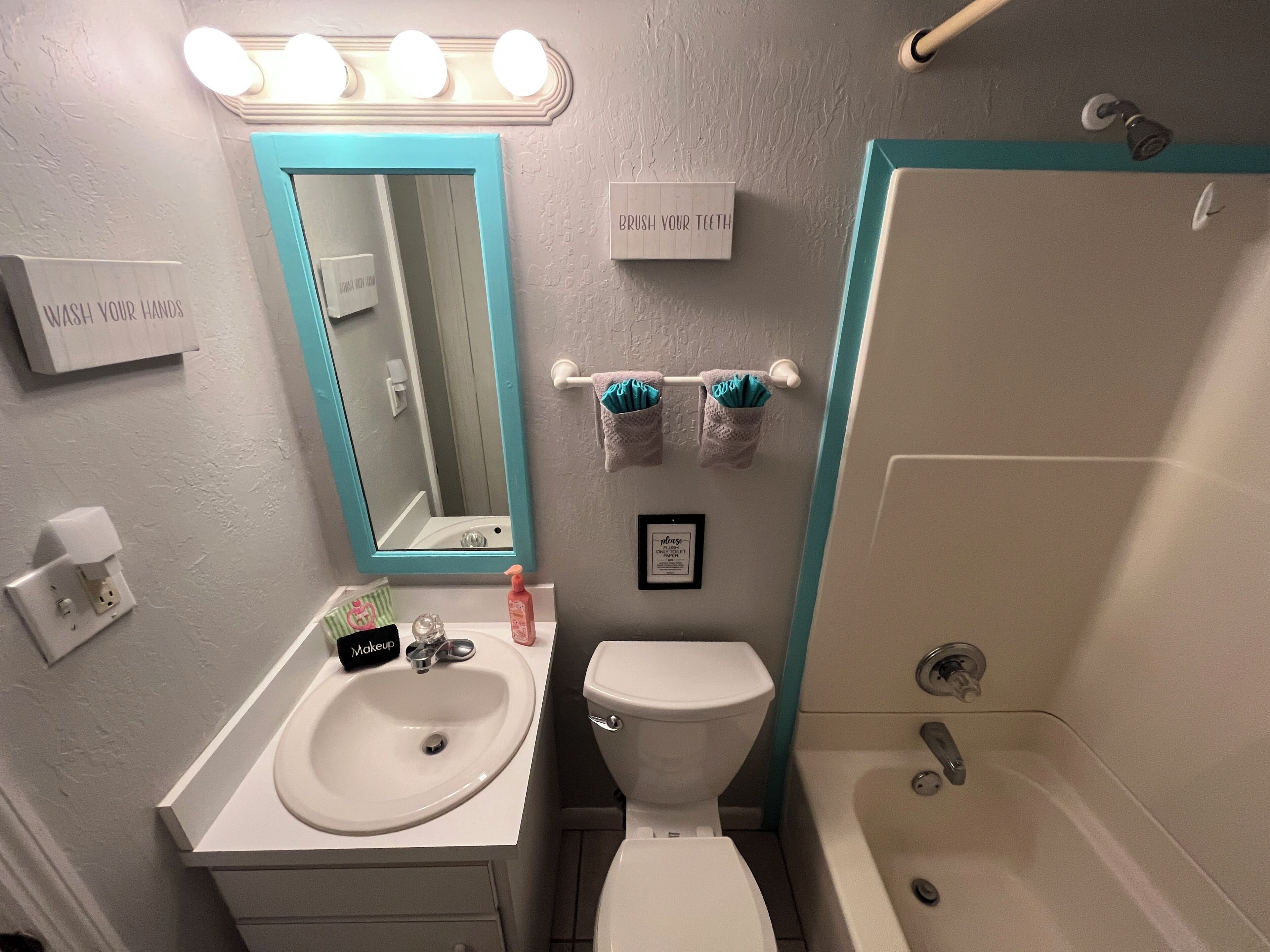 Un bagno con un lavandino bianco, uno specchio illuminato, un WC e una doccia. Sono presenti cartelli a parete e un portascopini con asciugamani.