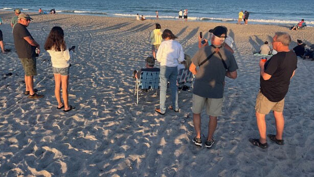 En sandstrand med folk som promenerar och sitter, ett lugnt hav och en klar himmel.