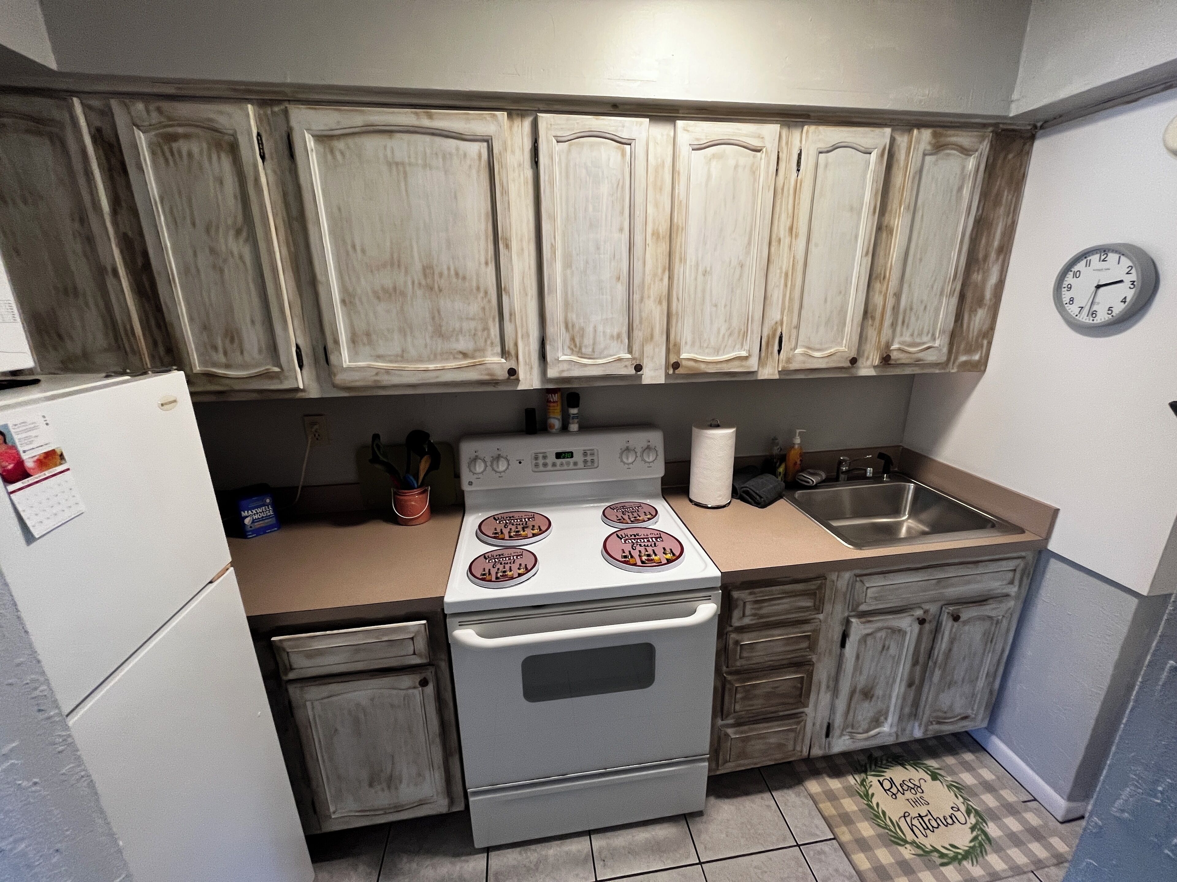 A compact kitchen with white cabinets, a stove, a sink, and a clock on the wall.