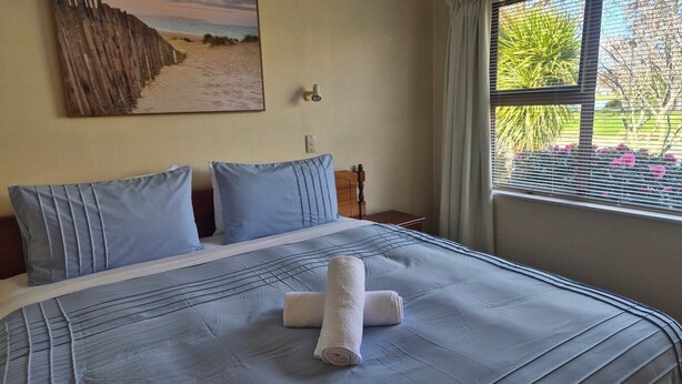 Ein ordentlich bezogenes Bett mit blauer Bettdecke und Kissen, ein Strandbild an der Wand und ein Fenster mit Blick auf Grünflächen und Blumen.