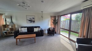 Ein Wohnzimmer mit einer schwarzen Couch, einem Holztisch, einem Bücherregal, einem Gemälde und einer großen Schiebeglastür zum Garten.
