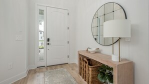 Un pasillo con una puerta blanca, un espejo redondo, una consola de madera con una lámpara y una cesta tejida.