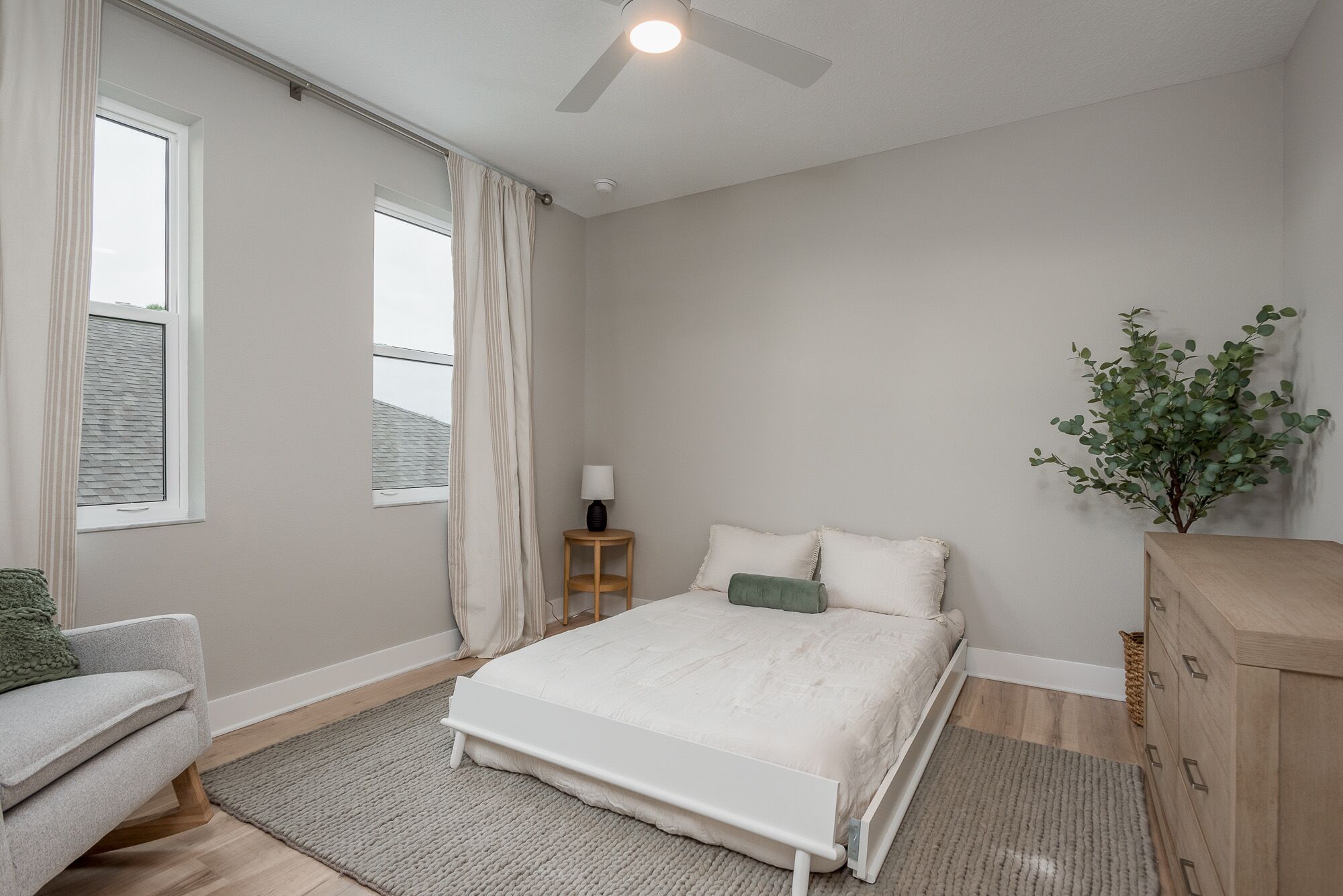 A bedroom with a bed, a nightstand, a lamp, a window with curtains, a plant, and a dresser.