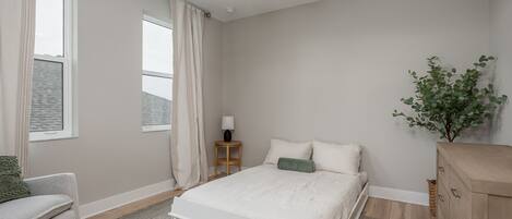 A bedroom with a bed, a nightstand, a lamp, a window with curtains, a plant, and a dresser.