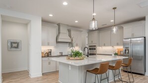 Una cocina moderna con armarios blancos, una isla central con taburetes de bar, electrodomésticos de acero inoxidable e iluminación colgante.
