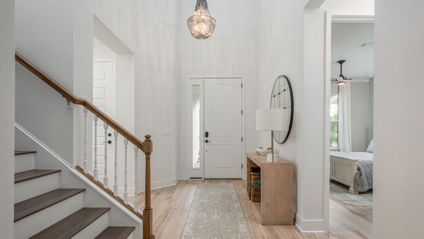 Un pasillo con una escalera de madera, un candelabro, una puerta, una lámpara y una cama al fondo.