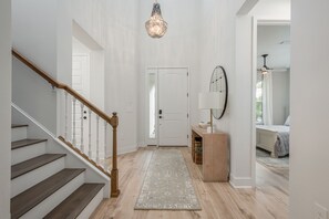 Un couloir avec un escalier en bois, un lustre, une porte, une lampe et un lit à l’arrière-plan.