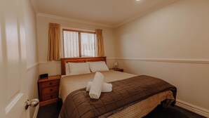 A neatly made bed with a brown quilt, a wooden nightstand with a lamp, and a window with curtains. - Alpine Springs Motel | Unit 7 - HS51217 (Hanmer Springs)