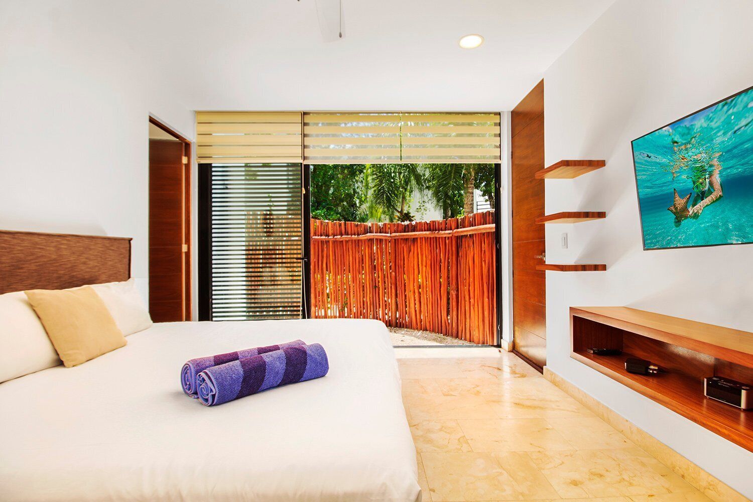 A modern bedroom with a large bed, a TV mounted on the wall, and a sliding glass door leading to a balcony.