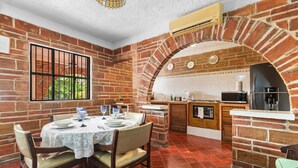 A dining area with a brick wall, a window with bars, a table set for two, and a kitchen area with a refrigerator and microwave.