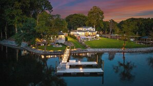 Una casa grande con un muelle que se adentra en un lago tranquilo, rodeada de exuberante vegetación y un césped bien cuidado.