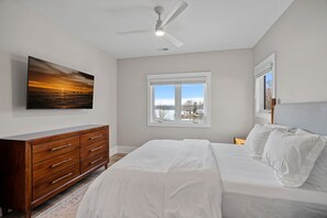 A bedroom with a bed, a dresser, a ceiling fan, a window with a view, and a painting on the wall. - Sunset Retreat at the Lake | Sleeps 24 | Pool & Hot Tub (Sherrills Ford)