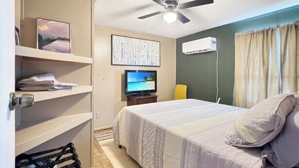 A hotel room with a bed, a television, a ceiling fan, an air conditioning unit, and a shelf with folded towels.