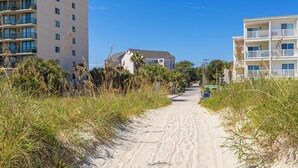 A sandy pathway flanked by tall grass and buildings with balconies, leading to a house with a sloped roof. - Beachside Bliss • Pool • Near Beach & Attractions (North Myrtle Beach)