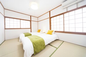 Perinteinen japanilaistyylinen huone, jossa on tatamimatto, kaksi sänkyä vihreillä ja keltaisilla vuodevaatteilla sekä suuret ikkunat puukarmeilla.