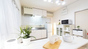 A modern kitchen with white cabinets, a microwave, a kettle, and a wooden cutting board. - Miraie Yokkaichi Aobacho (Yokkaichi)