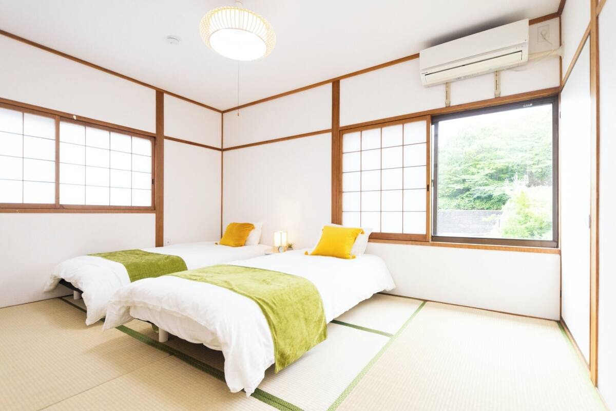 Um quarto tradicional japonês com tatames, duas camas com roupa de cama verde e amarela, uma janela grande com vista para vegetação e um aparelho de ar condicionado.