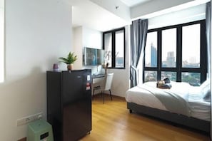 A modern hotel room with a bed, a desk, a refrigerator, and a window with a city view.