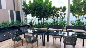Uma área de refeições moderna ao ar livre, com mesa de vidro e cadeiras de vime, rodeada por vegetação exuberante e vista para a cidade.