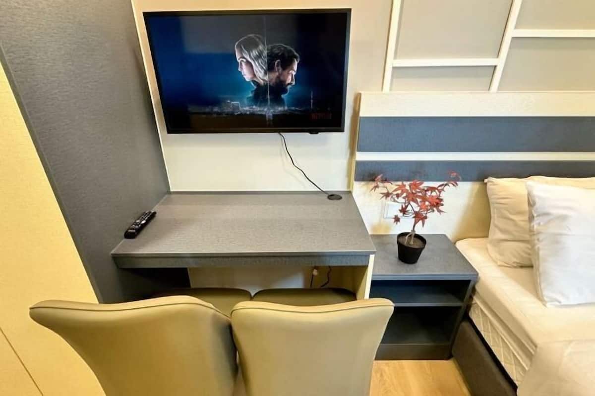 Een moderne hotelkamer met een flatscreen televisie aan de wand, een klein bureau met een afstandsbediening, een potplant en een bank.