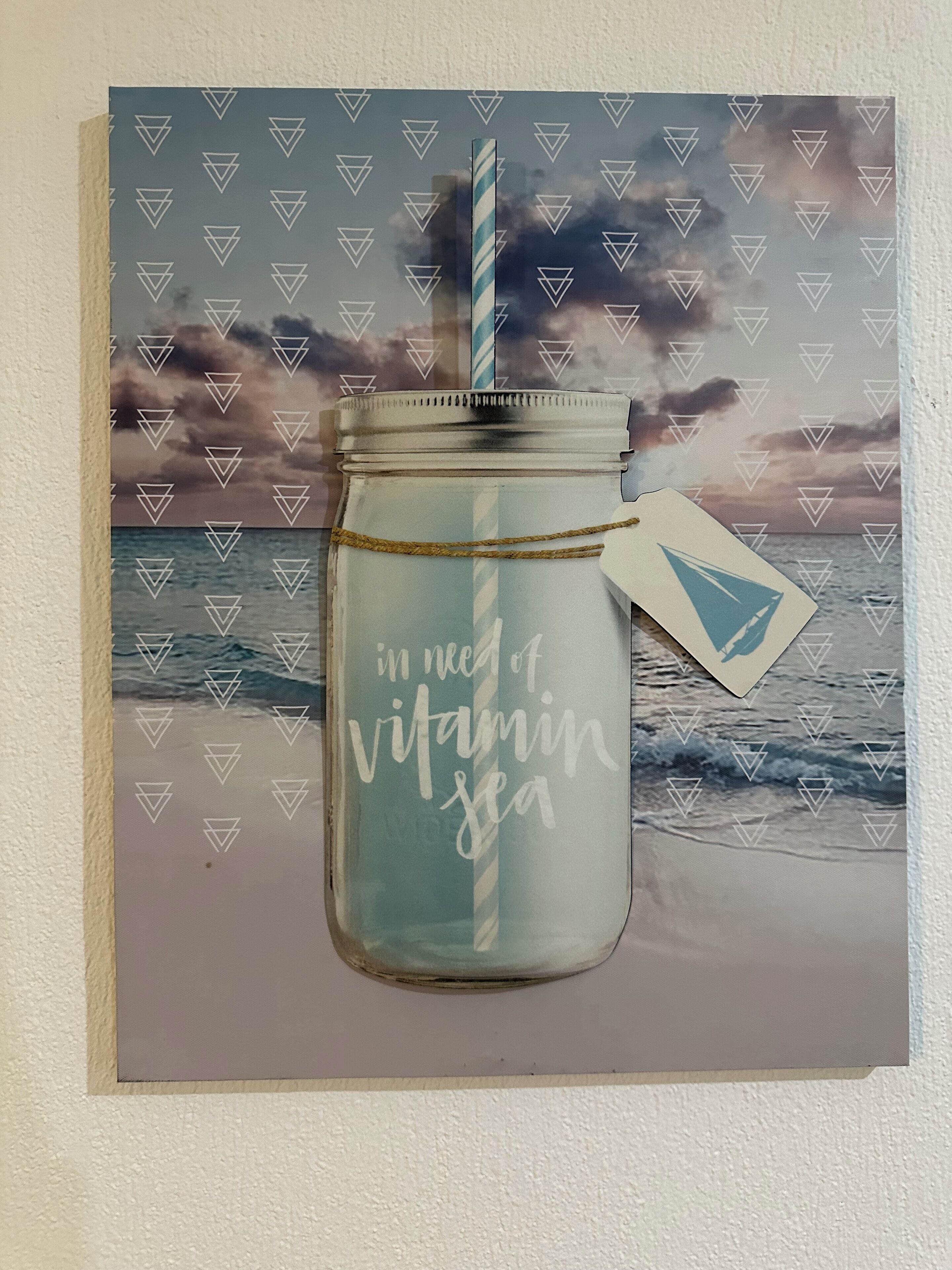 Una obra de arte enmarco muestra un tarro de cristal con una pajita y una etiqueta, con un paisaje marino de olas y un atardecer.