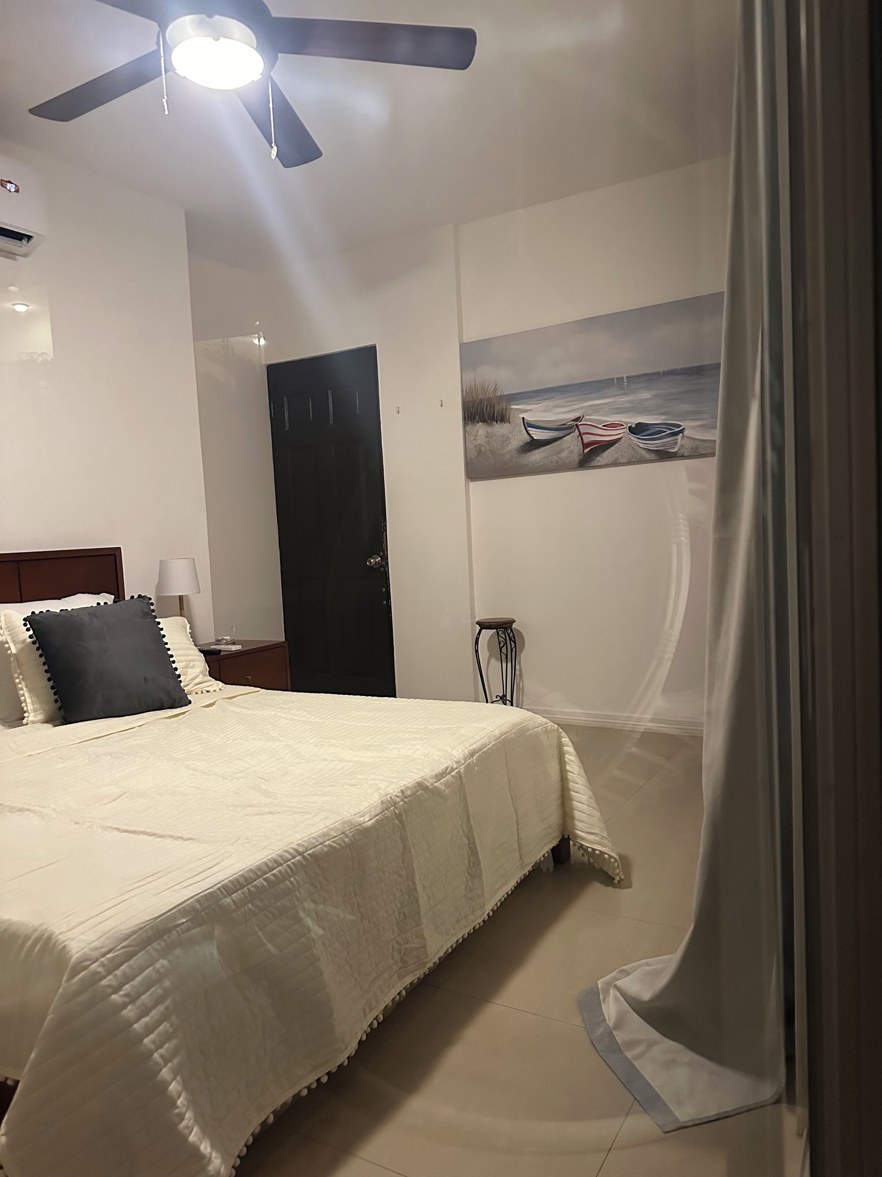 A bedroom with a bed, a ceiling fan, a painting of boats on the wall, and a door.