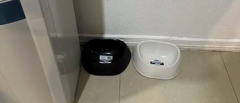 A corner of a room with a white wall and a tiled floor. There are two pet bowls, one black and one white, placed on the floor.