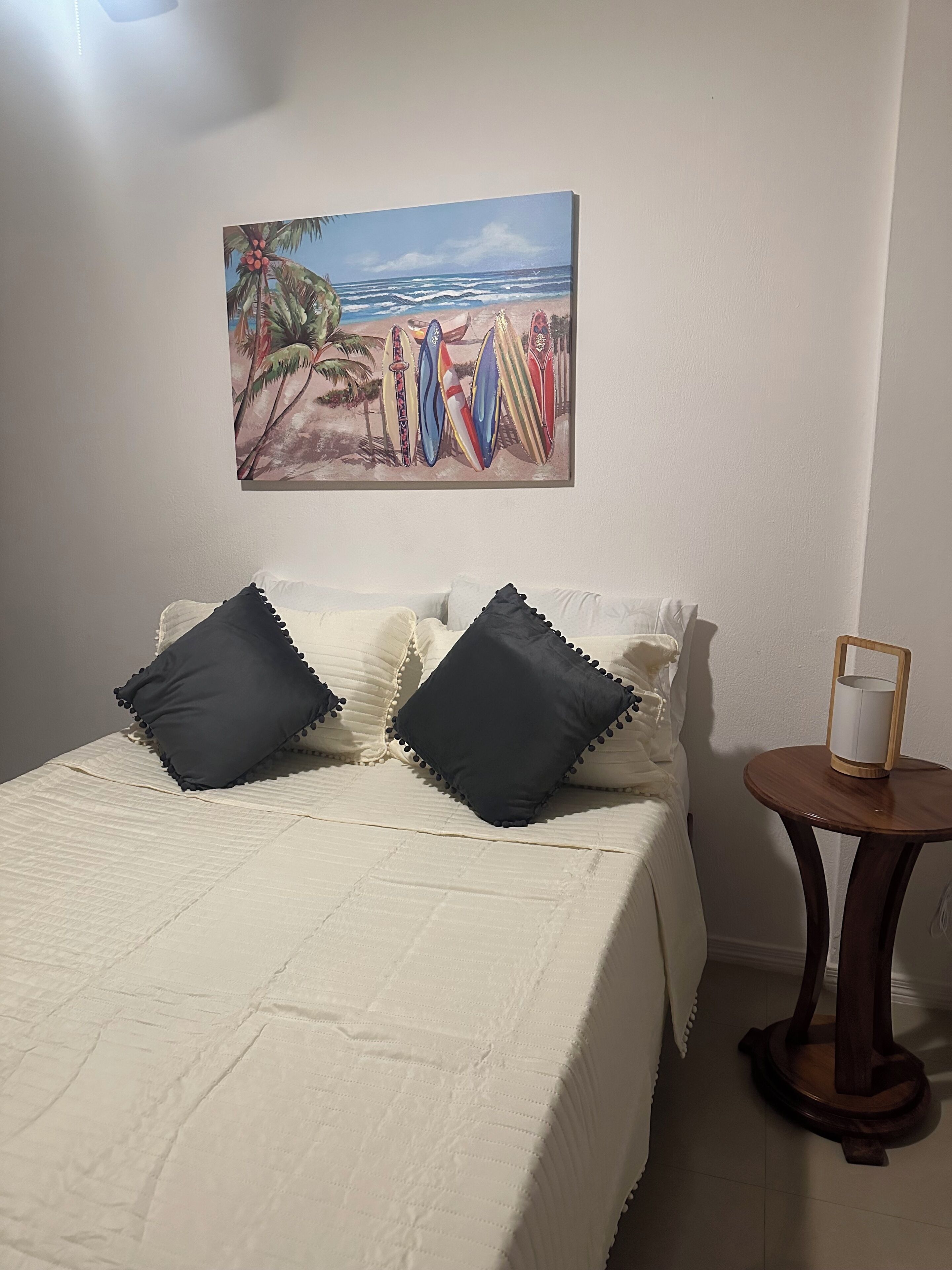 A neatly made bed with a white comforter and two dark pillows, a small wooden side table with a candle, and a beach-themed painting above the bed.