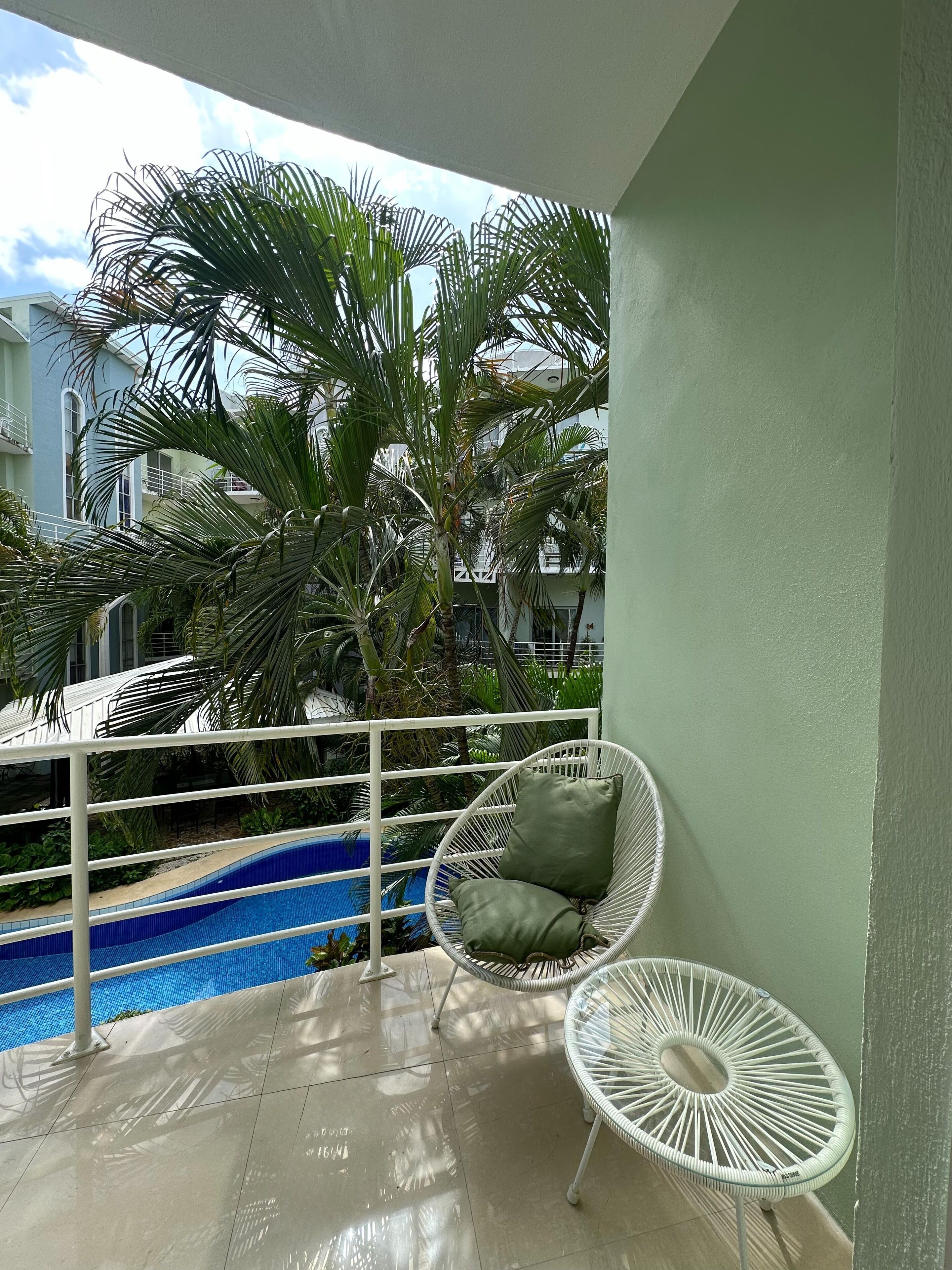 A balcony with a white chair and a small table, a pool, and a view of a building with balconies.