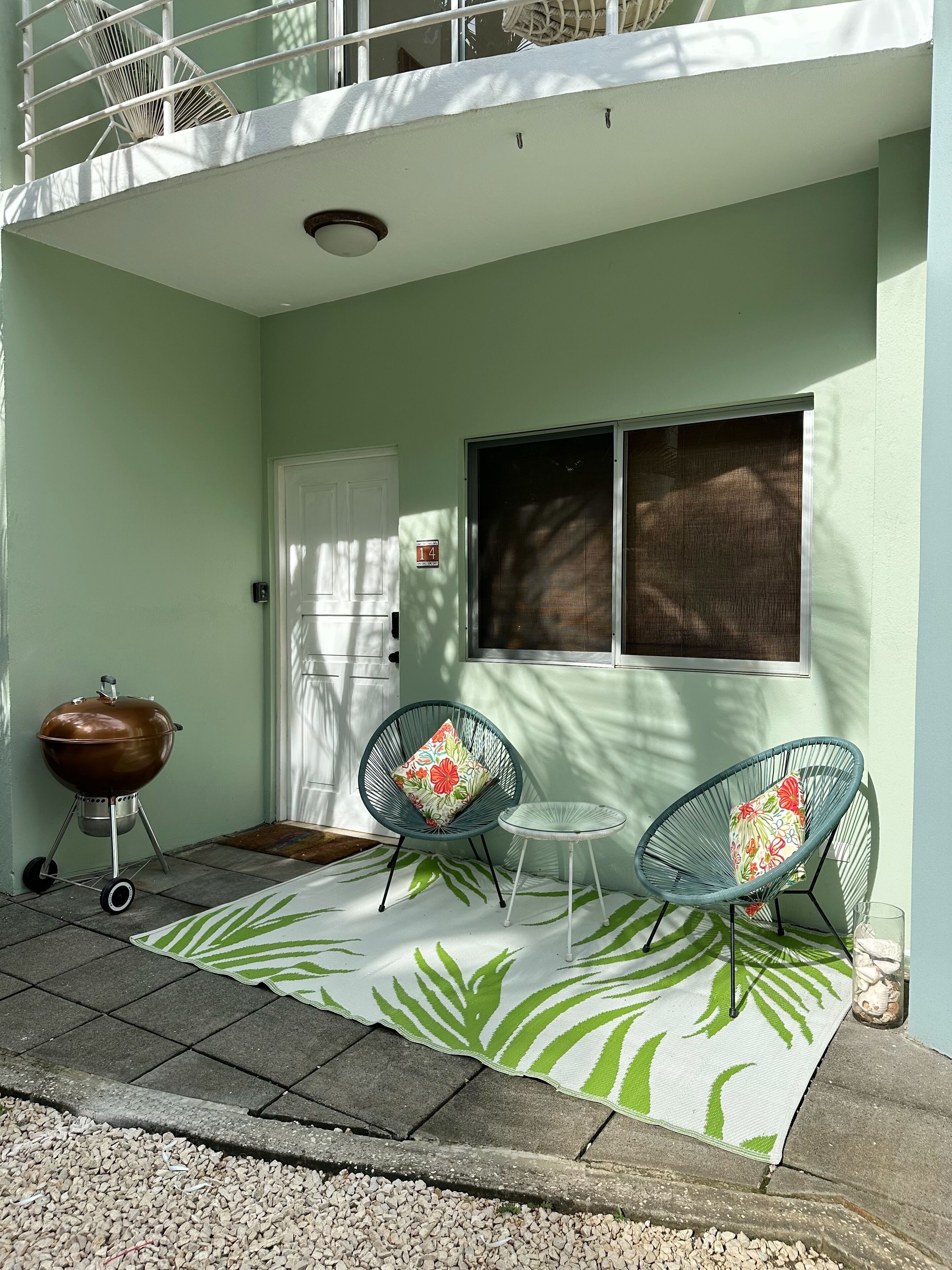 Área externa coberta com churrasqueira, duas cadeiras de vime, uma mesa pequena e um tapete estampado.