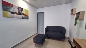 Uma sala de estar pequena e moderna, com um sofá escuro, um banquinho combinando e um quadro abstrato colorido na parede.