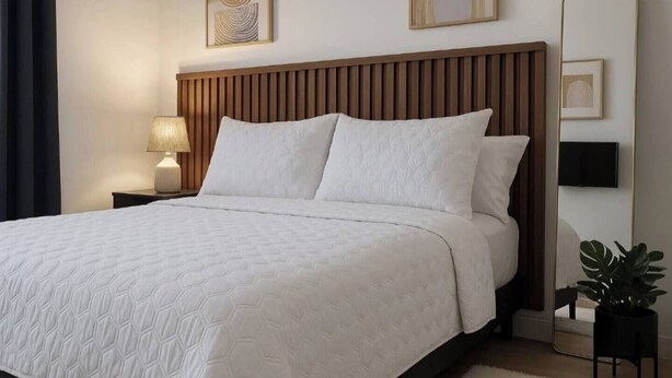 Cama bem arrumada com cabeceira de madeira, luminária de cabeceira e um espelho grande na parede.