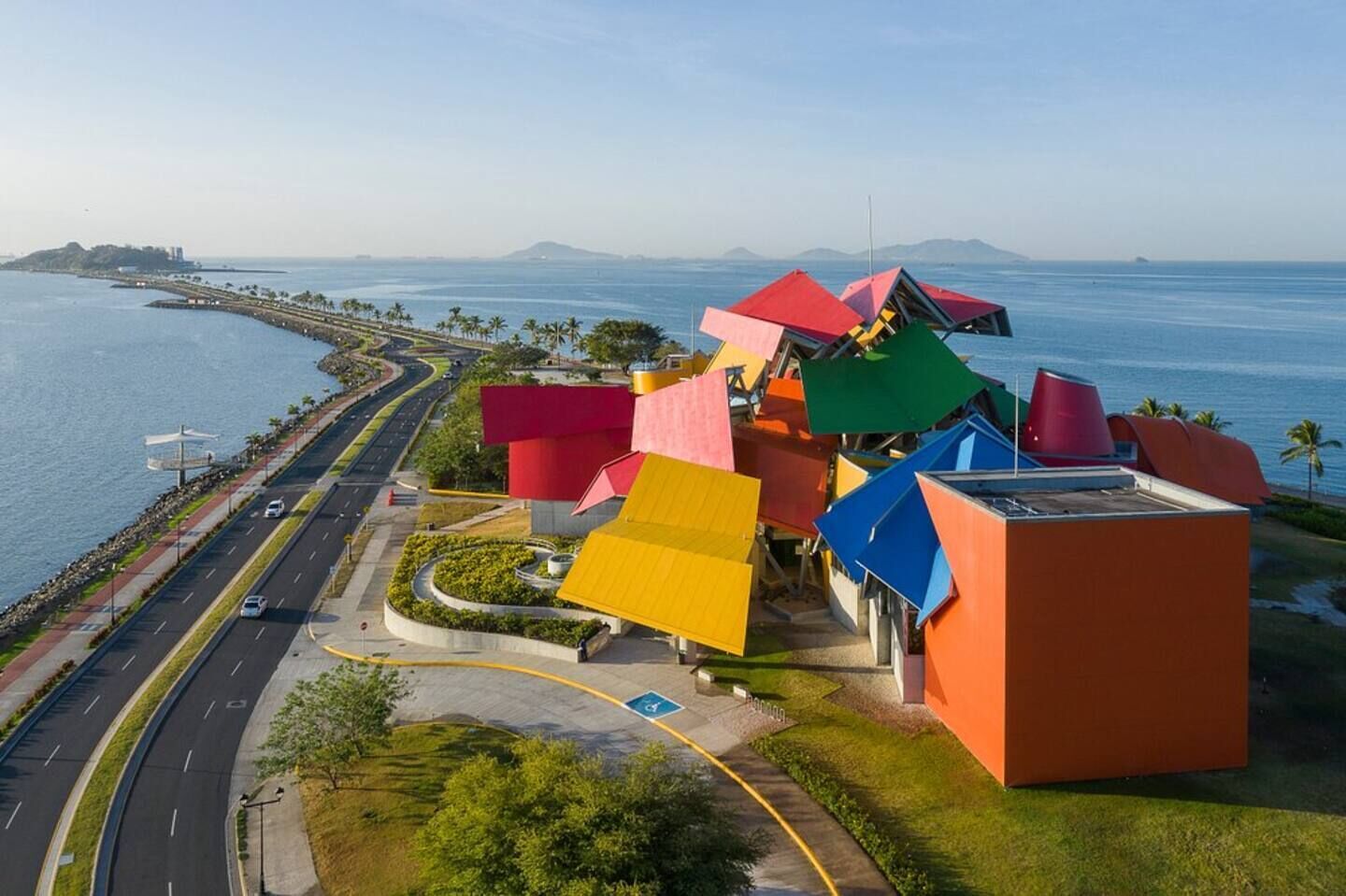 A modern building with colorful geometric shapes, a road with cars, and a body of water in the background.